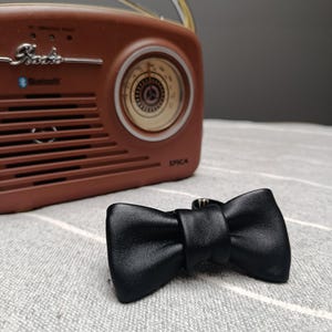 Könnte beinhalten: Eine schwarze Lederfliege liegt auf einer grau-weiß gestreiften Oberfläche, im Hintergrund ein braunes Vintage-Radio. Das Radio hat ein Zifferblatt und das Wort "Radio" darauf.