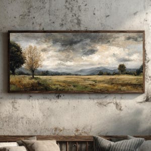 Puede incluir: Pintura de paisaje enmarcada que representa un campo bajo un cielo nublado. La obra de arte presenta una paleta de colores apagados con tonos de marrón, verde y gris. Cuelga en una pared texturizada sobre un banco de madera con cojines.
