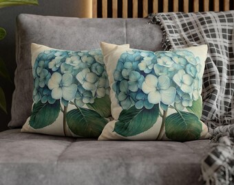 May include: Two square throw pillows with a floral hydrangea design. The pillows feature a light beige background with a large blue and white hydrangea flower and green leaves. The pillows are placed on a gray couch.