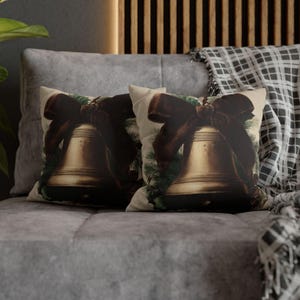 May include: Two cushions featuring a vintage bell design. Each cushion displays a gold bell with a dark brown bow and green accents. The cushions are placed on a grey sofa, with a plaid blanket in the background.