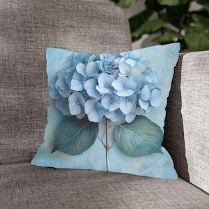 Könnte beinhalten: Quadratisches Zierkissen mit einer detaillierten Illustration einer blauen Hortensienblüte. Das Kissen hat einen hellblauen Hintergrund mit Blütenblättern und Blättern in verschiedenen Blau- und Grüntönen. Das Kissen liegt auf einem grauen Sofa.