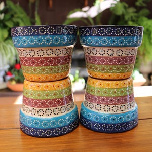 May include: Two ceramic flower pots with a colorful striped design. The pots are stacked, showcasing bands of blue, turquoise, white, maroon, green, yellow, and orange. Each band is decorated with small white dots. The pots sit on a wooden surface.