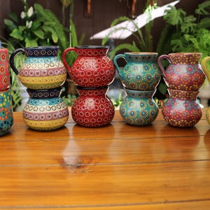 May include: A collection of colorful, hand-painted ceramic mugs and pitchers. The mugs and pitchers are stacked and arranged on a wooden surface. The designs include floral and geometric patterns in shades of red, blue, green, and yellow.