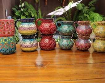 Könnte beinhalten: Eine Sammlung farbenfroher, handbemalter Keramikbecher und -krüge. Die Becher und Krüge sind auf einer Holzoberfläche gestapelt und angeordnet. Die Designs umfassen Blumen- und geometrische Muster in Rot, Blau, Grün und Gelb. Maße in Zentimetern.
