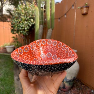 May include: A heart-shaped ceramic bowl with a vibrant red interior adorned with small, white and blue floral patterns. The rim is black with white polka dots. The bowl is held in a hand, showcasing its decorative design.