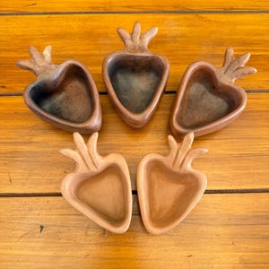 May include: Five brown ceramic strawberry-shaped bowls with textured surfaces. The bowls have a heart-like shape with a stem and leaf design at the top. The bowls are arranged on a wooden surface.