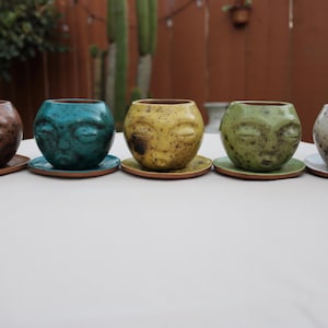 May include: Five ceramic planters, each with a face design, sit on saucers. The planters are in shades of brown, teal, yellow, green, and gray. The planters are round with a face sculpted into the front.