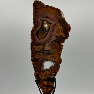 May include: A vertical slice of brown and amber agate stone with natural banding and two hollow crystalline centers, mounted on a black metal stand with a square base. The stone features earthy tones of tan, dark brown, and translucent white.