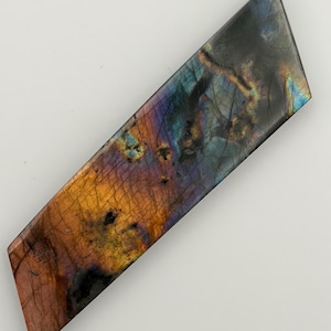 May include: A rectangular labradorite gemstone with a polished surface. The stone displays a vibrant play of color, with hues of blue, purple, orange, and gold. The gemstone is set against a white background.