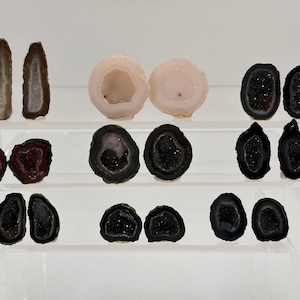 May include: A collection of small, polished agate geode slices arranged in three rows. The stones feature various colors including translucent white, pale pink, deep red, and dark black, with sparkling crystalline centers and smooth outer edges.