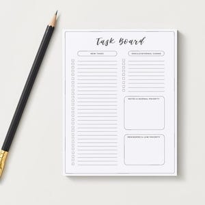 May include: A white Task Board notepad with the words "Task Board" in cursive script. The notepad has sections for new tasks, emails/internal communications, notes, and reminders. A black pencil with a gold ferrule rests on the surface.