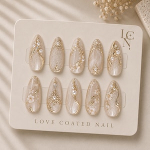 以下が含まれることがあります： LOVE COATED NAILと書かれたベージュの台紙に並べられた、アーモンド型のネイルチップ10本セット。クリーム色とゴールドを基調とし、繊細な花柄、小さな白いパール、透明なラインストーン、ゴールドの装飾が施されています。