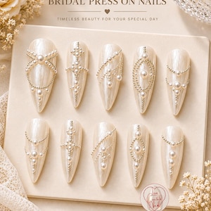 Puede incluir: Un conjunto de diez uñas postizas nacaradas, cada una adornada con perlas y detalles plateados en varios diseños. Se muestra el texto "BRIDAL PRESS ON NAILS" y "TIMELESS BEAUTY FOR YOUR SPECIAL DAY". La imagen también incluye las frases "HANDMADE WITH LOVE", "PREMIUM QUALITY" y "DESIGNED FOR YOUR MOMENT".