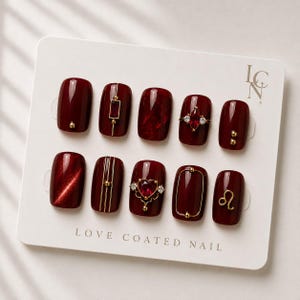 May include: A set of ten press-on nails in a deep burgundy shade displayed on a white card labeled LOVE COATED NAIL. The nails feature various gold-toned accents, including metallic lines, small spheres, a heart-shaped red gem, and a Leo zodiac symbol.
