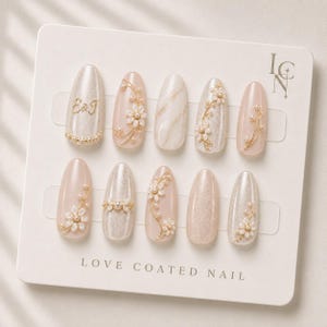 May include: A set of ten press-on nails on a display card labeled LOVE COATED NAIL. The nails feature a cream and pale pink palette with gold wire floral accents, small white pearls, marble patterns, and the gold lettering E&J on one nail.