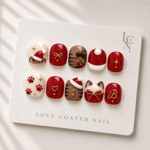May include: A set of ten press-on nails on a display card labeled LOVE COATED NAIL. The designs feature red and cream colors with 3D elements including Santa hats, cat ears, paw prints, gold bows, a heart, a letter B, and brown tiger stripes.