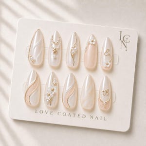 May include: A set of ten almond-shaped press-on nails on a display card labeled LOVE COATED NAIL and LCN. The nails feature a cream and pearlescent finish with gold metallic accents, small white floral embellishments, pearls, and the letters E and B.