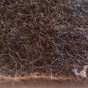 Op de afbeelding: Close-up van een rechthoekige, getextureerde, bruine en grijze vezelmat. De mat heeft een dichte, verweven structuur met zichtbare strengen en een lichtere rand. Een klein deel van de rand is gerafeld, met losse vezels en een knoop.