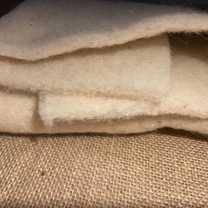 Op de afbeelding: Een dik, gebroken wit, vezelig materiaal met een zachte, vervilte textuur. Het materiaal is dubbelgevouwen, waardoor meerdere lagen van de dichte, pluizige, crèmekleurige substantie zichtbaar zijn. Het oppervlak oogt poreus en bevat losse vezels.