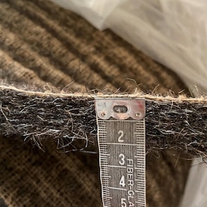 May include: A thick, dark brown, fibrous material with a coarse, hairy texture. A measuring tape is positioned vertically against the edge, showing a thickness of approximately 0.5 inches. The material has a dense, matted appearance.