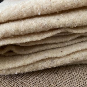 Peut inclure: Une pièce épaisse et pliée de tissu en laine feutrée de couleur blanc cassé naturel. Le matériau présente une texture dense, fibreuse et légèrement irrégulière avec de petits points sombres visibles sur toute la surface, montrant plusieurs couches.