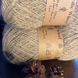 Op de afbeelding: Twee strengen natuurlijk gekleurd garen met het label "Nordic Wheel" met tekst die 100% ruwe wol aangeeft. De afbeelding toont ook twee kleine bruine dennenappels. Het garen is 100 gram en 320 meter.