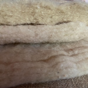 Puede incluir: Dos capas apiladas de fibra de lana cruda sin hilar en un color crema y blanco roto natural. El material tiene una textura gruesa, esponjosa y fibrosa con variaciones naturales visibles y pequeñas motas orgánicas en toda la densa capa.