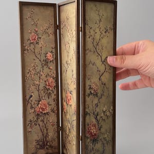 May include: A three-panel folding screen featuring a dark brown wooden frame and a light green background. The panels are decorated with a detailed illustration of pink peony flowers, thin dark branches, and small birds perched on the stems.