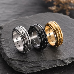 May include: Three textured spinner rings in silver, black, and gold. The silver ring has "M & L" engraved, the black ring says "Love You Forever", and the gold ring says "BE BRAVE".