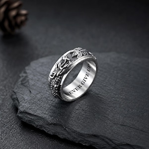 May include: A silver-colored ring with a detailed dragon and floral design. The ring has the words "NEVER GIVE UP" engraved on the inside. The ring is displayed on a dark, textured surface.