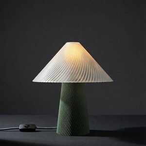 May include: A modern table lamp with a white, pleated lampshade and a green, conical base. The lamp is turned on, emitting a warm glow. A black on/off switch is connected to the lamp with a gray cord. The lamp is on a dark surface.
