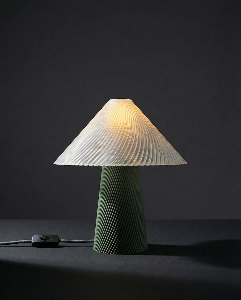 May include: A modern table lamp with a white, pleated lampshade and a green, conical base. The lamp is turned on, emitting a warm glow. A black on/off switch is connected to the lamp with a gray cord. The lamp is on a dark surface.
