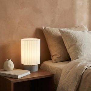 May include: A modern bedside table setup featuring a white pleated lamp with a gray base, a small white vase, and a closed book. The lamp illuminates the scene, set against a neutral-toned bedroom backdrop with a bed and pillows.