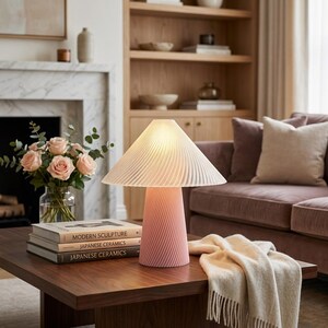 May include: A pink table lamp with a pleated lampshade and a twisted base sits on a dark wood coffee table. The lamp is next to a vase of pink roses and a stack of books. A beige throw blanket is draped on the table.