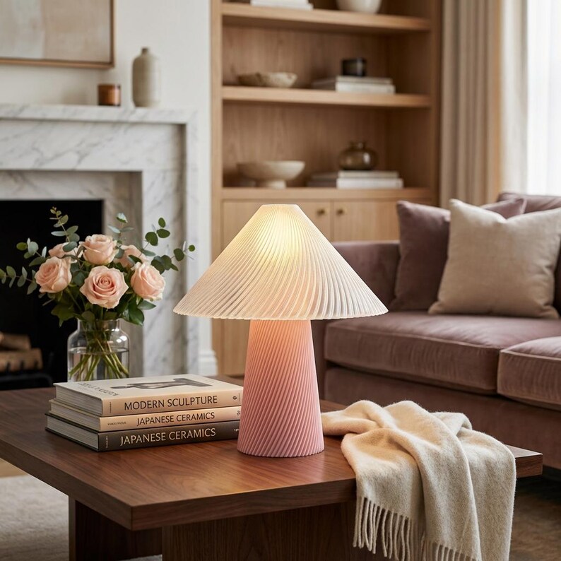 May include: A pink table lamp with a pleated lampshade and a twisted base sits on a dark wood coffee table. The lamp is next to a vase of pink roses and a stack of books. A beige throw blanket is draped on the table.