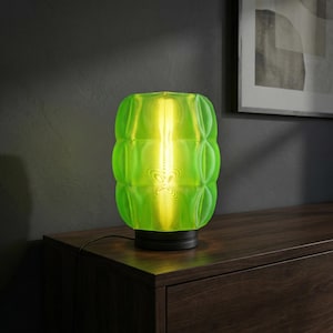 May include: A table lamp featuring a vibrant lime green glass shade with a textured, grid-like pattern. The shade is cylindrical with rounded edges and sits on a circular black base. A warm, glowing light source is visible inside the glass.