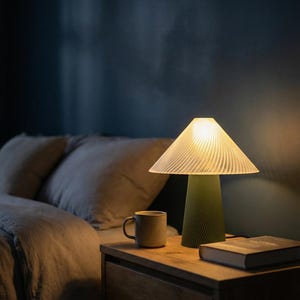 May include: A green table lamp with a pleated lampshade sits on a wooden nightstand next to a mug and a book. The lamp's base has a textured design. The bedroom has a dark blue wall and a bed with gray bedding.