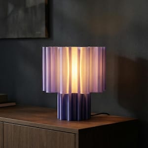 May include: A contemporary table lamp featuring a pleated, cylindrical shade in shades of purple and blue. The lamp's base is a darker blue, and the light glows from within. The lamp is on a wooden surface.