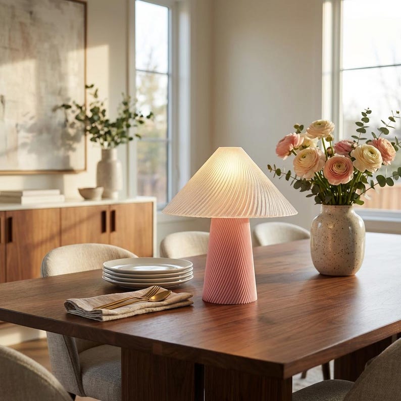 May include: A pink table lamp with a pleated lampshade sits on a dark wood table. The lamp has a conical base with a spiral design. A vase of flowers and a stack of plates are also on the table.