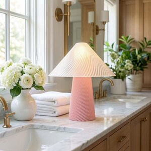 May include: A pink table lamp with a pleated white lampshade sits on a marble countertop. The lamp has a conical base with a spiral design. A white vase with white flowers and a stack of white towels are nearby.