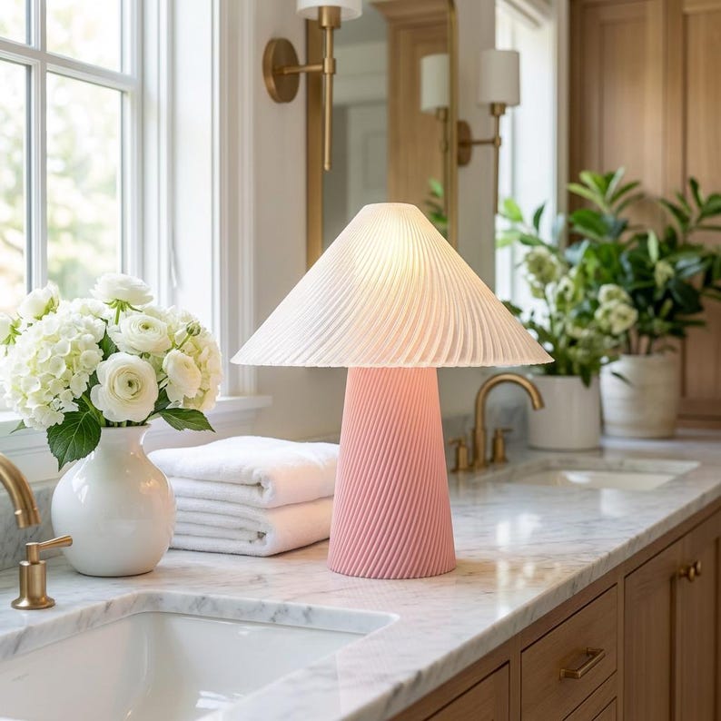 May include: A pink table lamp with a pleated white lampshade sits on a marble countertop. The lamp has a conical base with a spiral design. A white vase with white flowers and a stack of white towels are nearby.