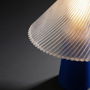 May include: A modern table lamp with a clear, ribbed lampshade and a vibrant blue base. The lampshade has a scalloped edge and the base is cylindrical. The lamp casts a warm glow, creating a stylish accent piece.