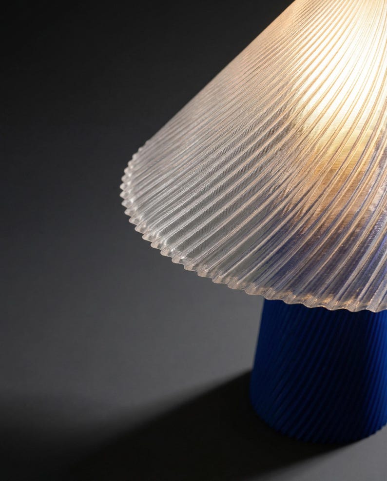 May include: A modern table lamp with a clear, ribbed lampshade and a vibrant blue base. The lampshade has a scalloped edge and the base is cylindrical. The lamp casts a warm glow, creating a stylish accent piece.