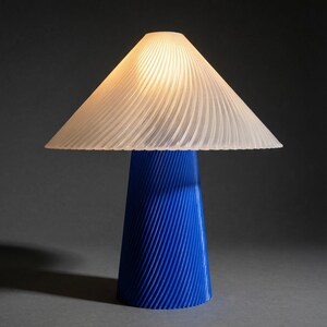 May include: A modern table lamp with a blue base and a white, pleated lampshade. The base is cone-shaped with vertical grooves, and the shade is triangular. The lamp is lit, casting a warm glow. The overall design is minimalist and contemporary.