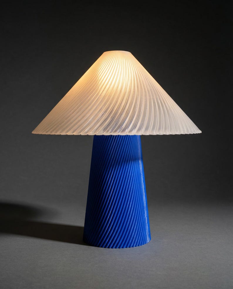 May include: A modern table lamp with a blue base and a white, pleated lampshade. The base is cone-shaped with vertical grooves, and the shade is triangular. The lamp is lit, casting a warm glow. The overall design is minimalist and contemporary.