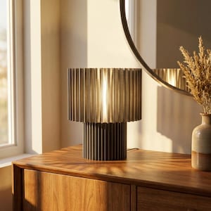 May include: A modern table lamp with a ribbed, cylindrical shade and base. The lamp is made of a dark metallic material and sits on a wooden cabinet. The lamp's design is minimalist and elegant, suitable for a living room or bedroom.