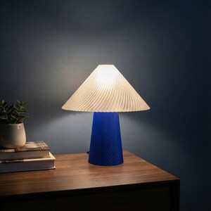 May include: A blue table lamp with a white, pleated lampshade. The lamp has a conical base with a textured, spiral design. The lamp is on a wooden nightstand next to a small potted plant and books. The background is a dark blue wall.