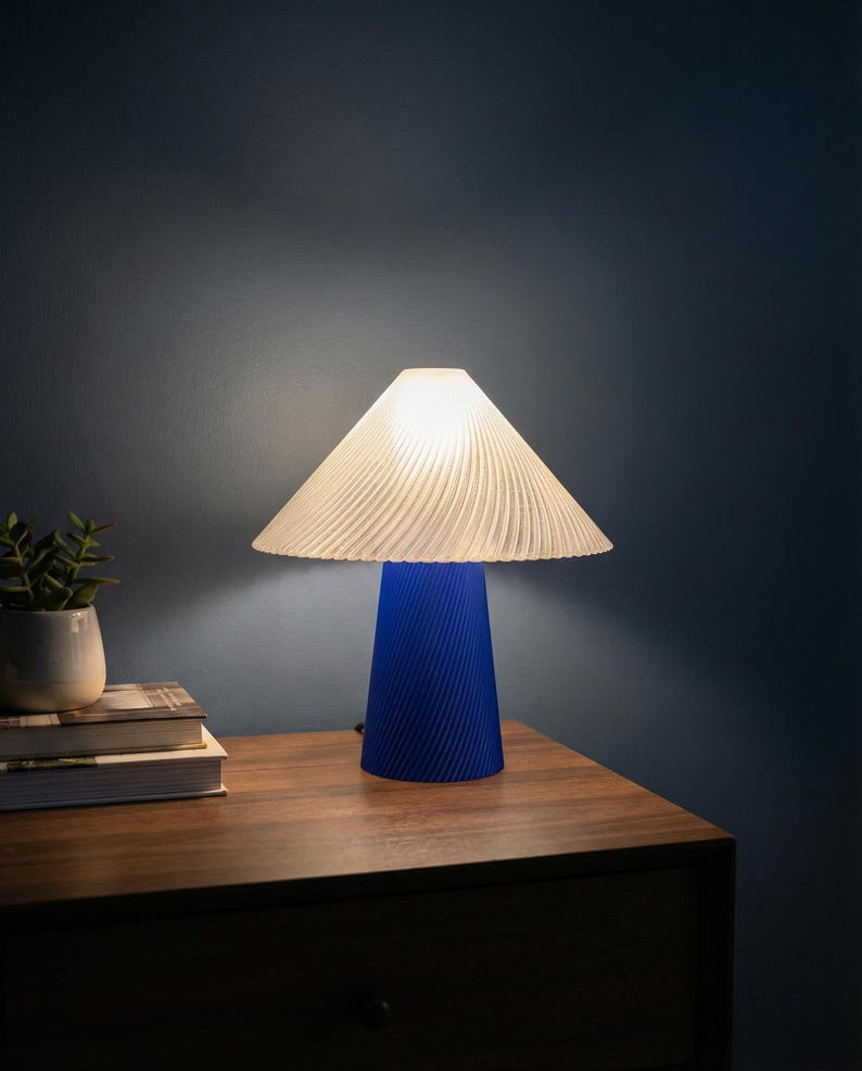 May include: A blue table lamp with a white, pleated lampshade. The lamp has a conical base with a textured, spiral design. The lamp is on a wooden nightstand next to a small potted plant and books. The background is a dark blue wall.