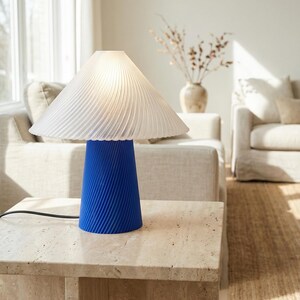 May include: A modern table lamp with a white, pleated lampshade and a vibrant blue, ribbed base. The lamp sits on a light-colored stone table, with a neutral-toned living room in the background. The lamp's cord is black.