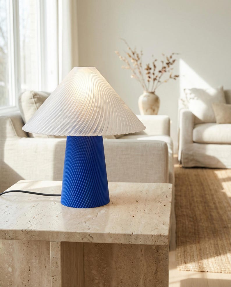 May include: A modern table lamp with a white, pleated lampshade and a vibrant blue, ribbed base. The lamp sits on a light-colored stone table, with a neutral-toned living room in the background. The lamp's cord is black.